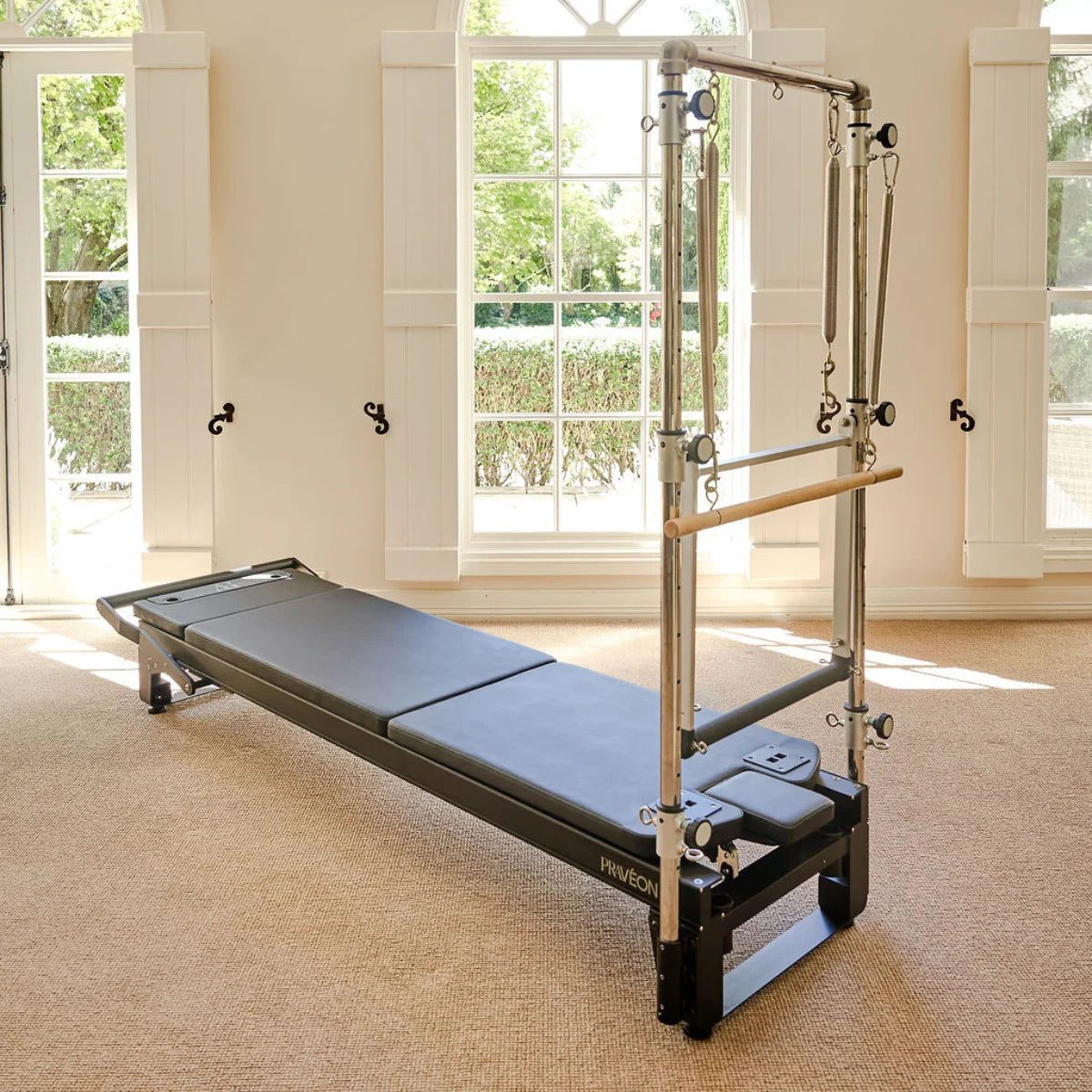 Professional PRAVEON Pilates Cadillac Reformer with a black padded carriage and stainless steel tower frame in a sunlit studio.