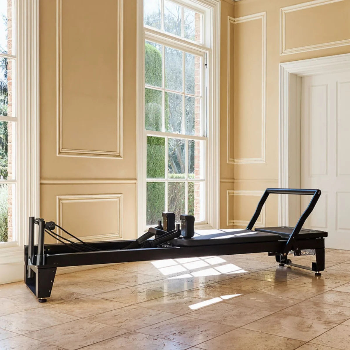 Black professional Pilates reformer machine positioned on a tiled floor in a sunlit room with large windows and classic wall moulding.