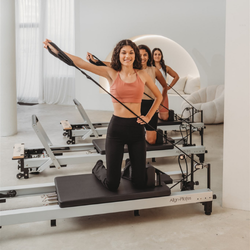 Woman using a Pilates reformer machine with another person in the background.