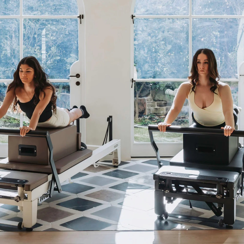 Two women using Pilates reformers in a bright, modern studio with large windows and a tiled floor.