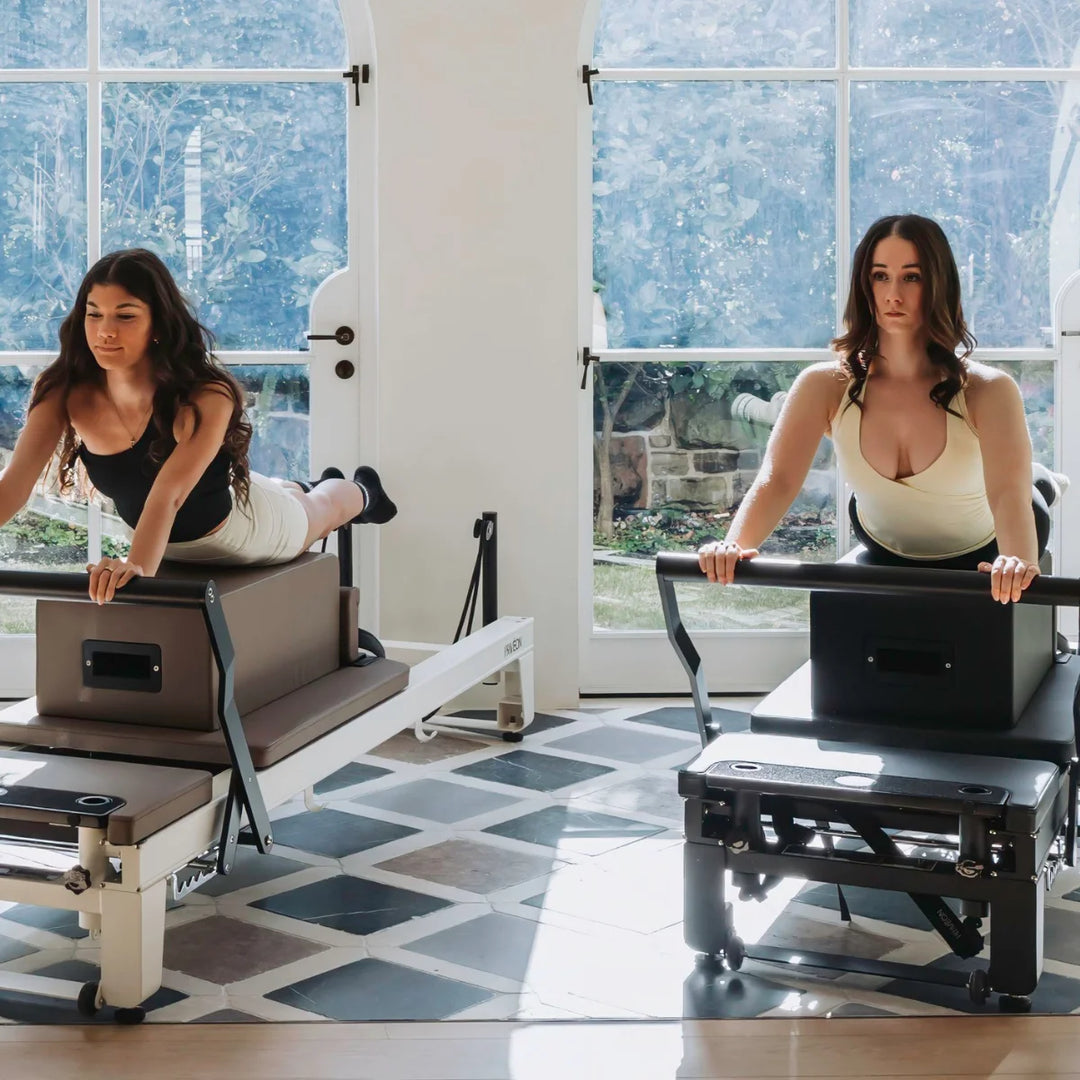 Two women using Pilates reformers in a bright, modern studio with large windows and a tiled floor.
