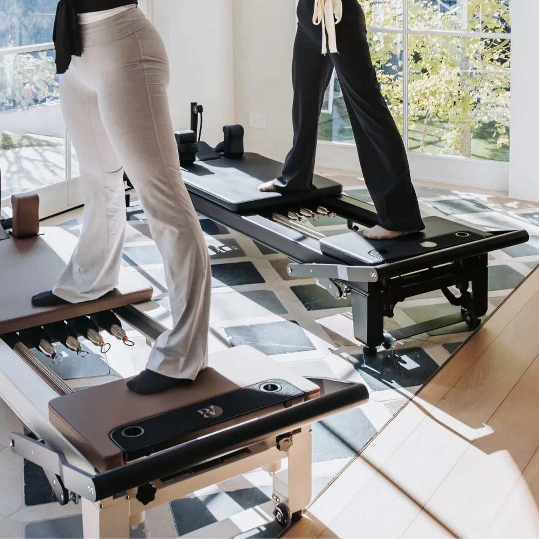 Two people using Pilates reformer platform extender in a bright, sunlit studio with large windows and a geometric rug.