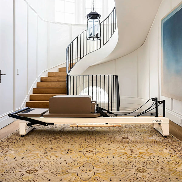 A Pilates reformer machine with a ivory aluminium frame and brown carriage, positioned on a patterned yellow rug in front of a white spiral staircase with wooden steps and a black metal railing.