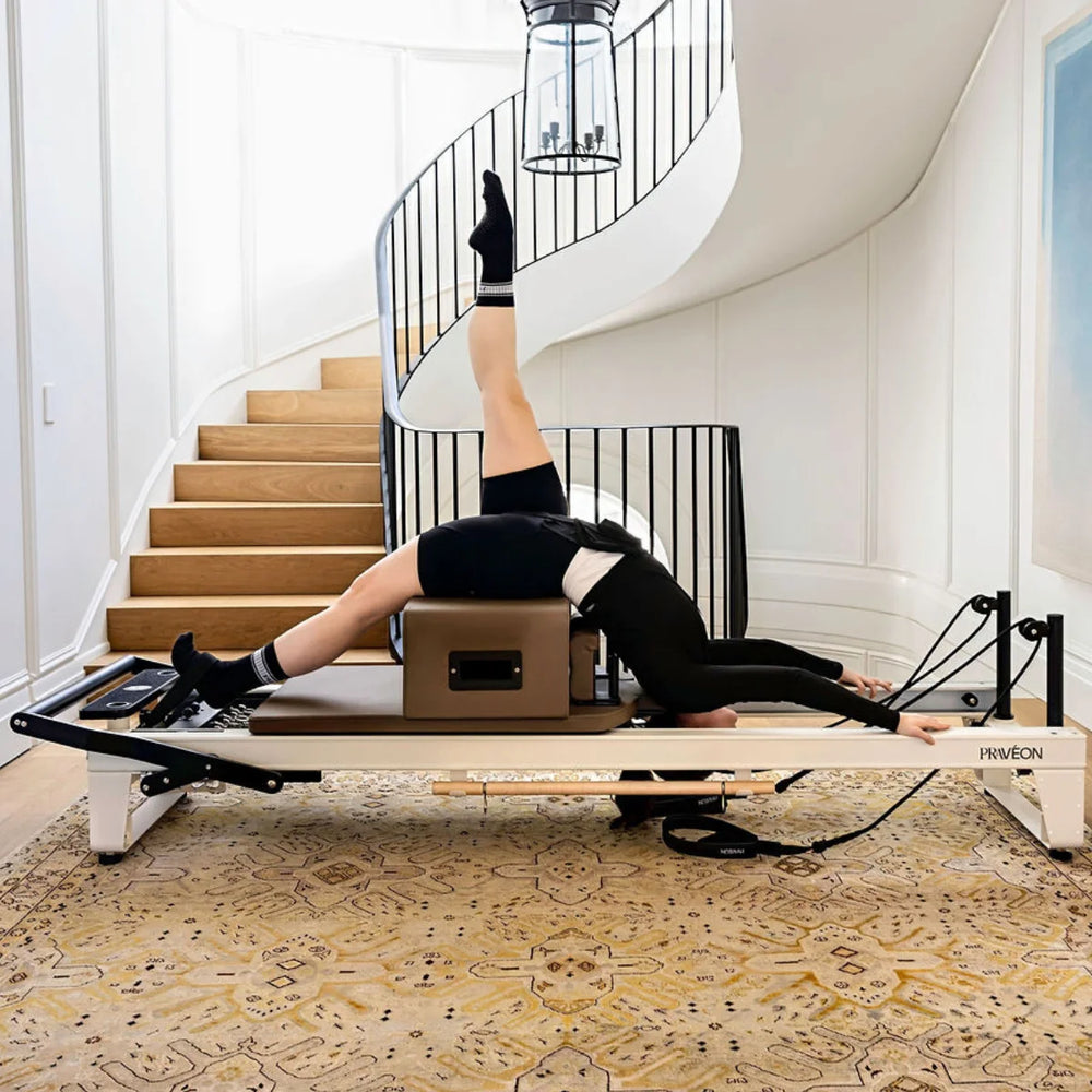 A person in black activewear performs a pilates exercise on a Pilates reformer machine in a bright, modern home with a white spiral staircase and patterned beige rug.