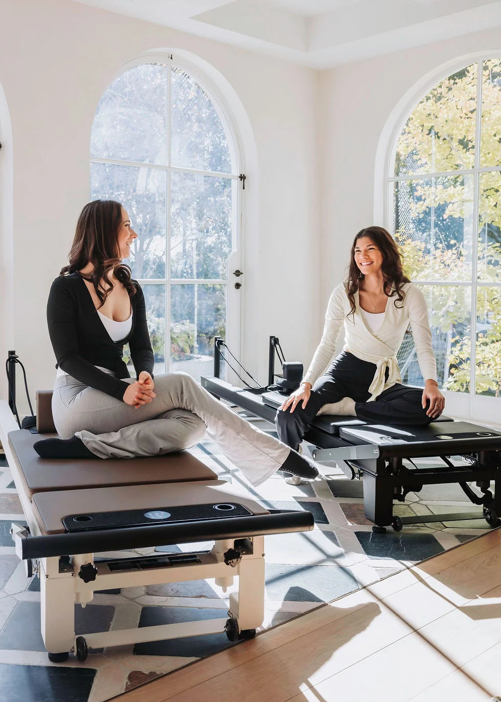 Two women sitting on Pilates reformers in a bright room with large windows.