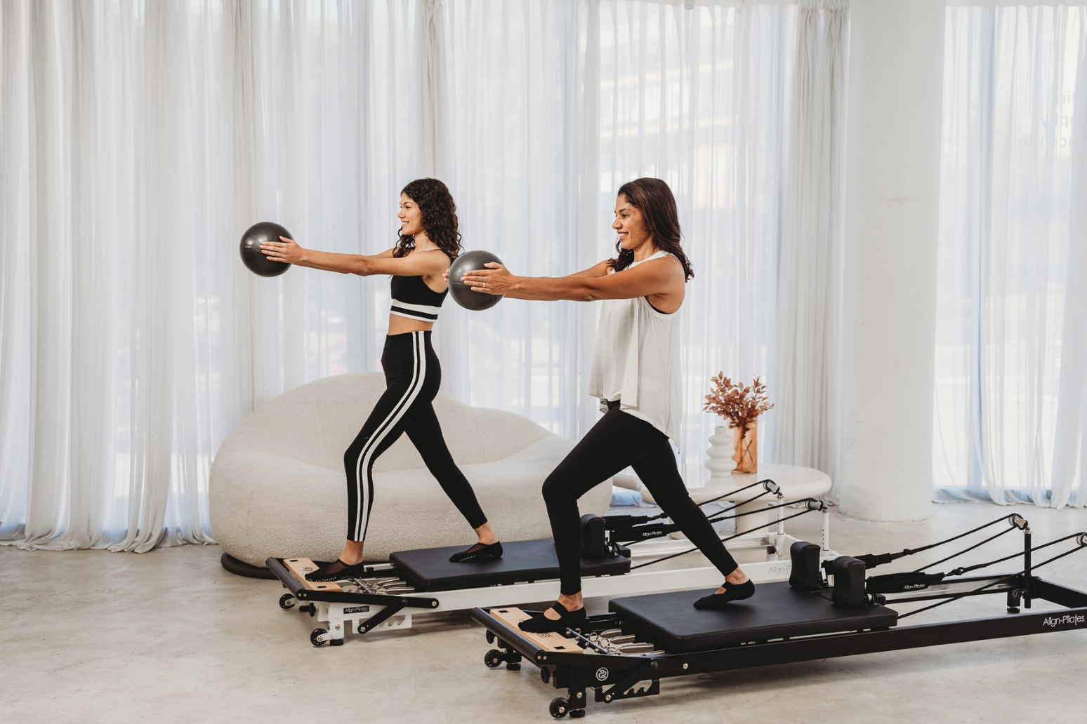 Two women exercising on Pilates reformers in a bright room with white curtains.