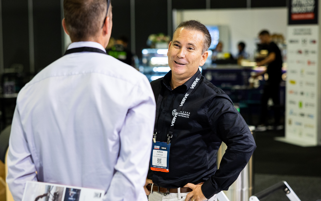 Two men engaged in a conversation at an event with booths and displays in the background.