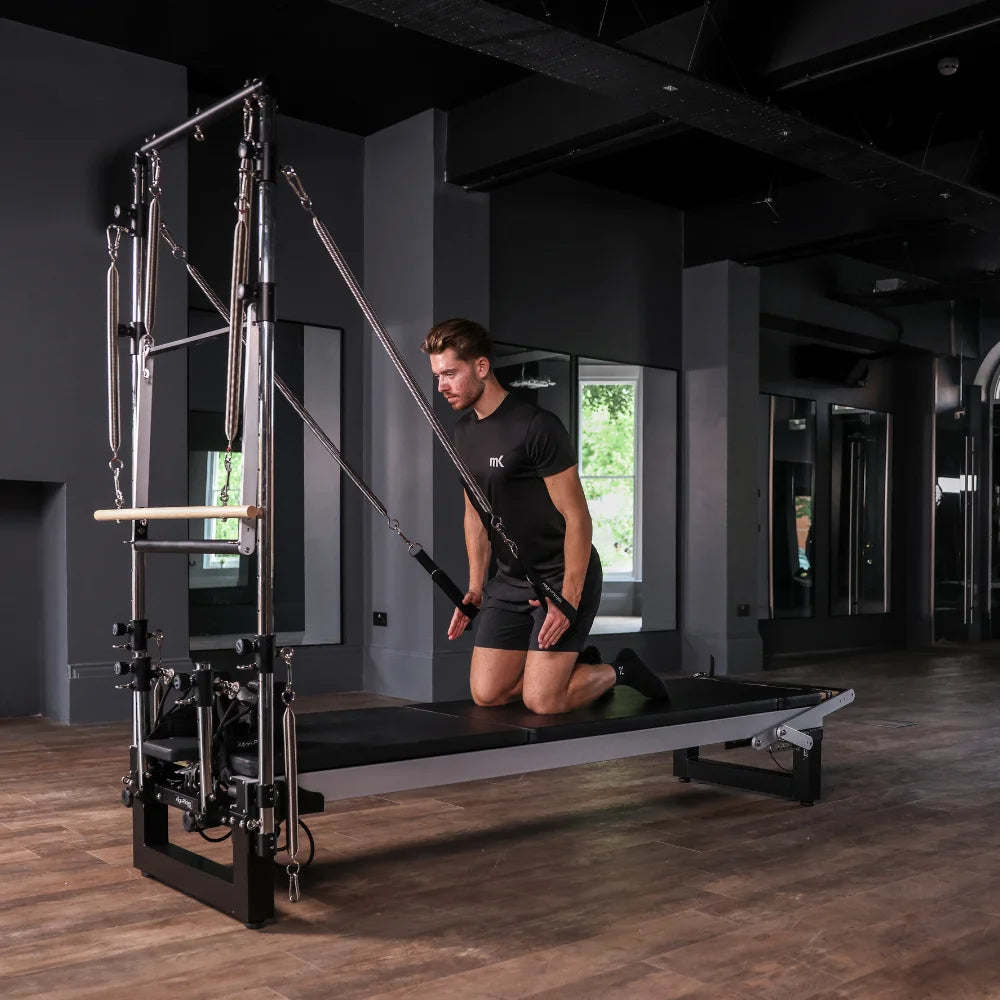 Person using a Pilates reformer machine with half trapeze in a dark room with wooden flooring.