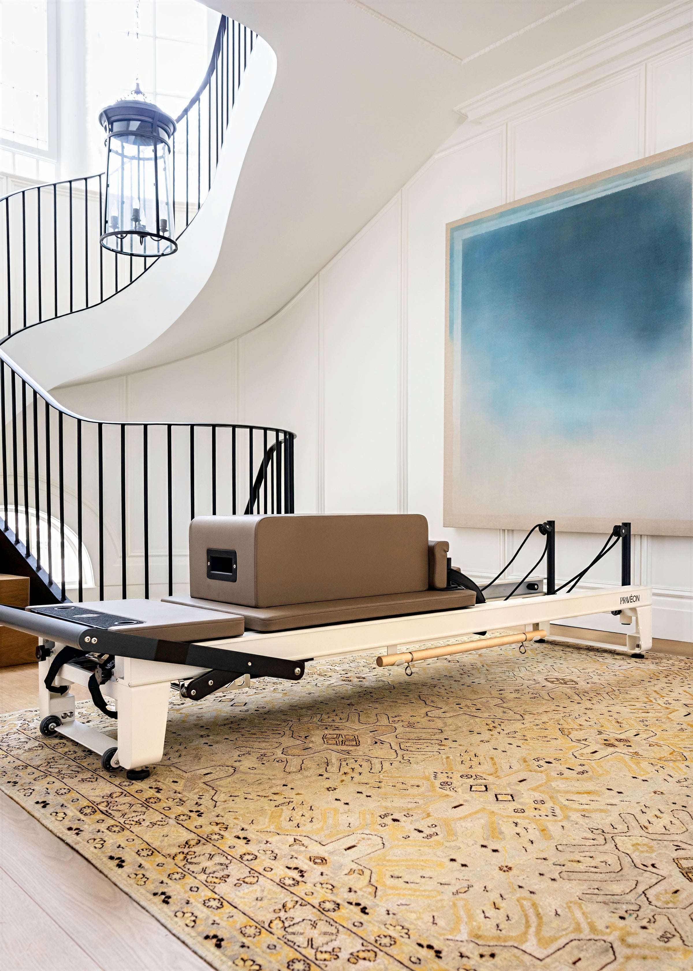A Pilates reformer machine with a taupe-brown padded carriage and white frame, positioned on a patterned beige rug in a bright room with a white spiral staircase and a large blue-toned abstract painting on the wall.