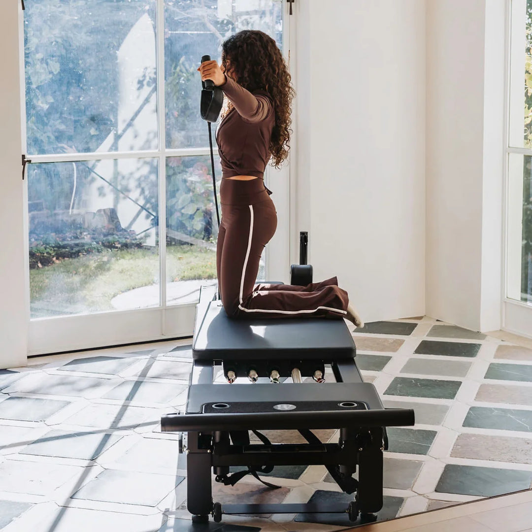 A woman in a matching brown activewear set kneels on a Pilates reformer machine in a bright, modern room with large windows and tiled floors.