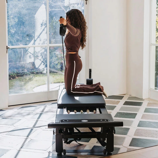A woman in a matching brown activewear set kneels on a Pilates reformer machine in a bright, modern room with large windows and tiled floors.