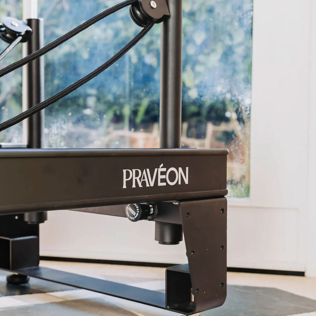 Close-up of a black PRAVÉON reformer exercise machine with the brand name printed in white on the side, set in a bright room with a window in the background.