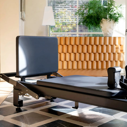 A black Pilates reformer machine photographed indoors in a modern room with a wood-paneled wall, window, lamp, and tiled floor.